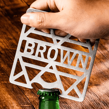 Hand using a stainless steel coaster with 'BROWN' cutout as a bottle opener on a green bottle, wooden background.