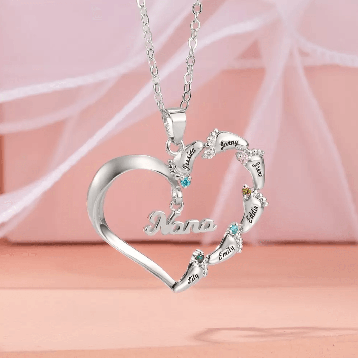 A silver heart-shaped necklace with "Nana" inscribed, featuring eight names (Lily, Emily, Eddie, etc.) adorned with colorful gemstones, displayed against a soft pink background.