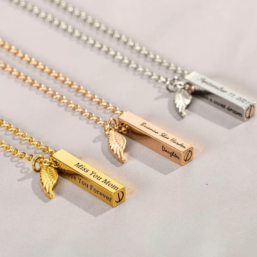 Gold and silver engraved bar urn necklaces with feather charms, featuring personalized messages laid on a soft surface