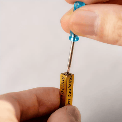 Hands using a tool to secure the screw on a gold bar urn necklace, engraved with 'Miss You Mom' and a date