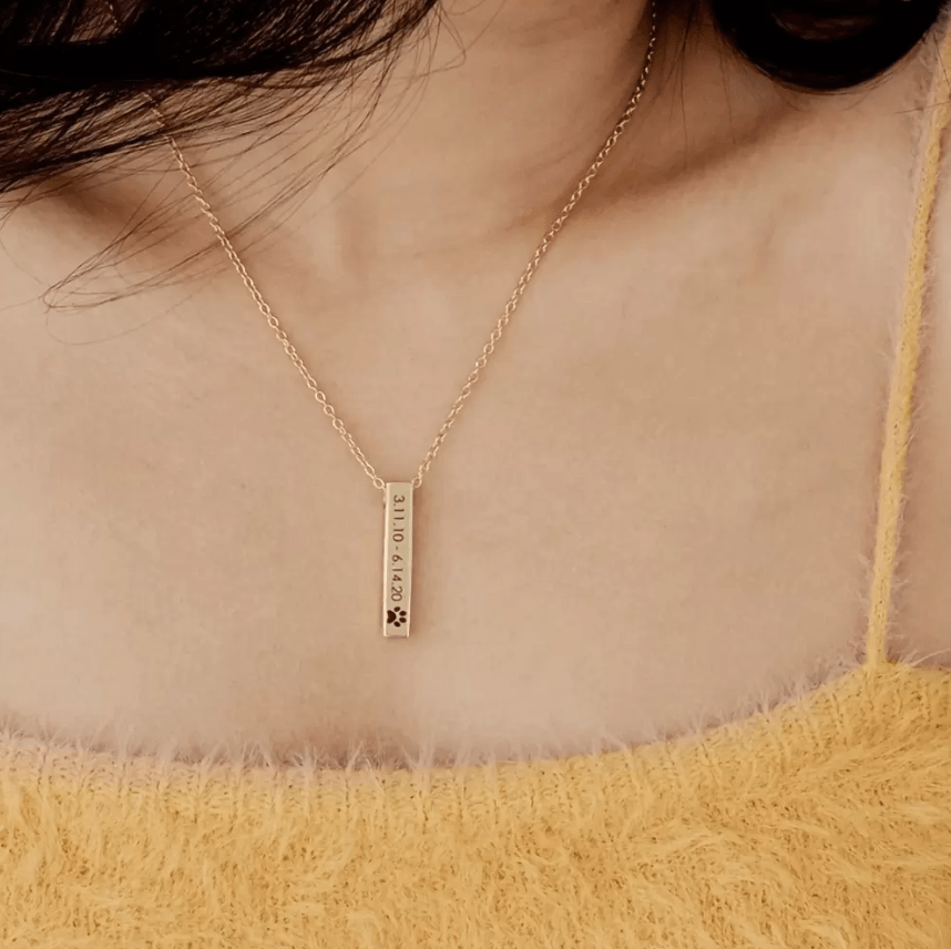 Gold bar urn necklace worn by a woman, engraved with symbolic icons, displayed on a yellow sweater