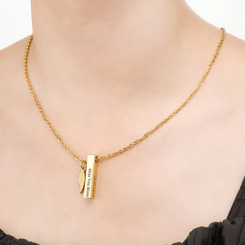 Close-up of a gold bar urn necklace with an engraved message and feather charm, worn on a delicate chain