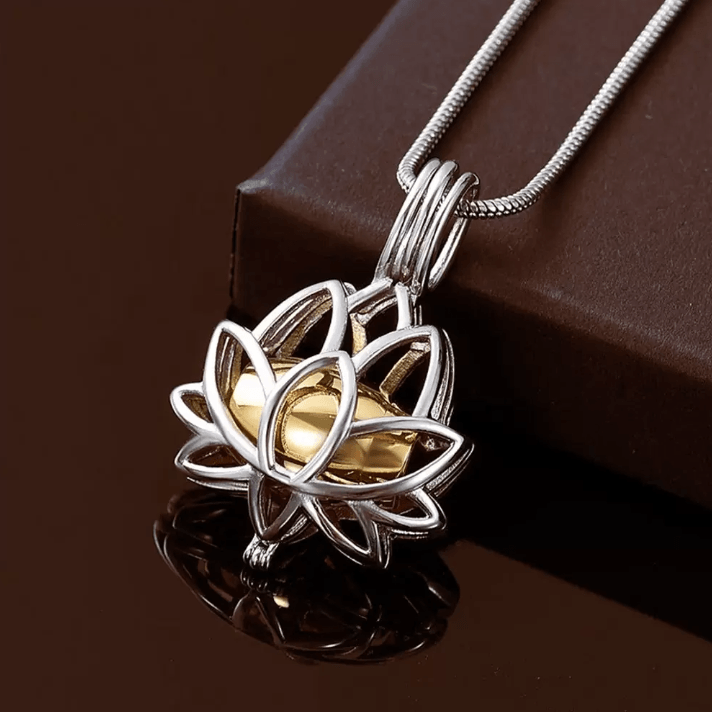 Silver lotus cremation urn necklace with a golden center, reflecting on a dark surface beside a box.