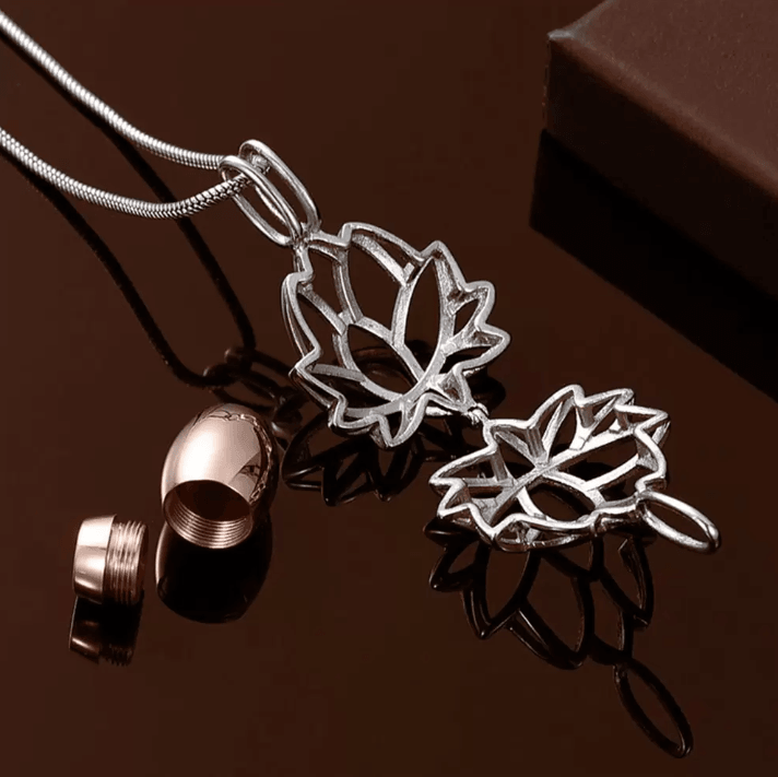 Rose gold and silver lotus cremation urn necklaces with an open capsule, on a reflective brown surface.