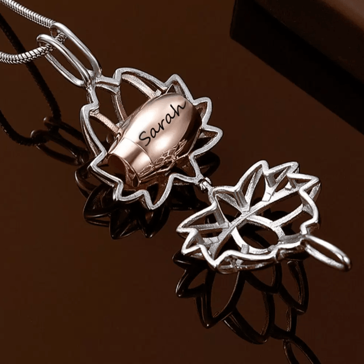 Rose gold cremation urn necklace with 'Sarah' engraving, shaped as a lotus flower, displayed on a reflective surface.