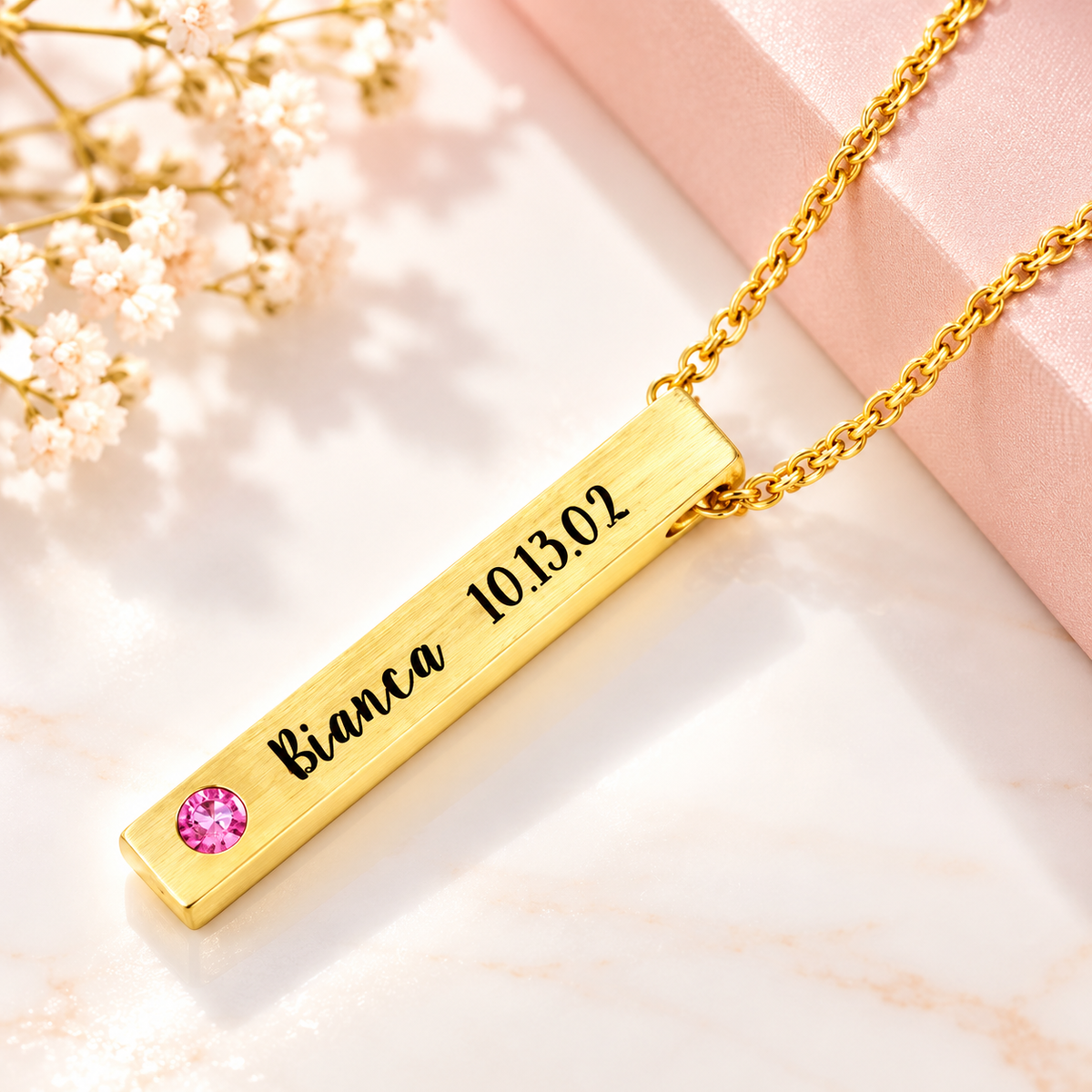 Personalized gold-tone 4-sided bar necklace with brushed finish, Bianca name and date engraving, and pink birthstone, displayed on cream marble with delicate flowers.