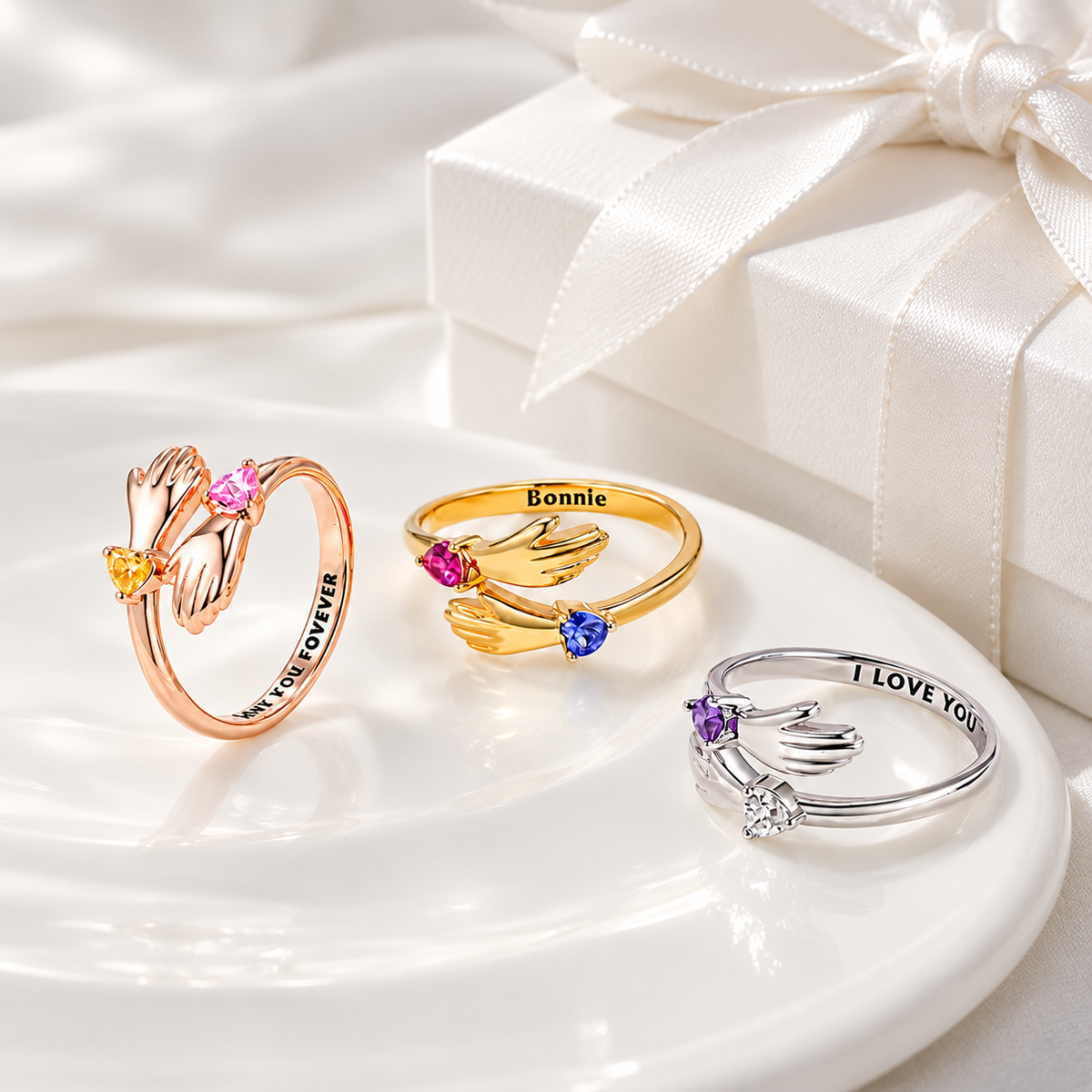 Personalised hug rings in rose gold, gold and silver with heart-shaped birthstones and engraved messages, displayed beside a white gift box. 