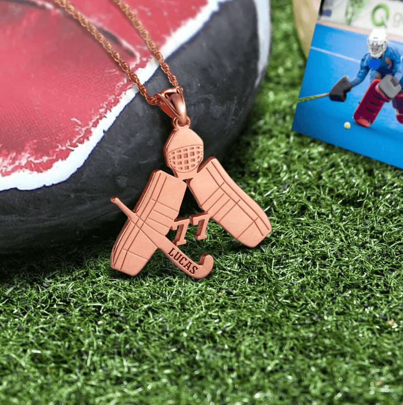 A rose gold hockey-themed pendant with a goalie mask, crossed goalie pads, a stick, and the number 17 on grass, with a photo of a goalie in the background.