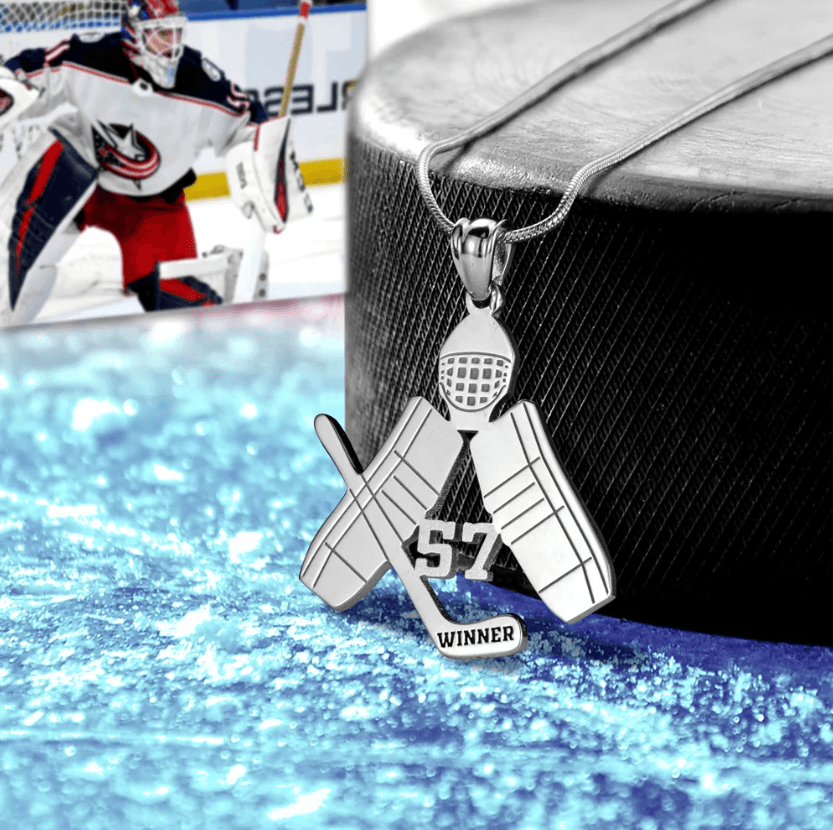 A silver hockey-themed pendant with a goalie mask, crossed goalie pads, a stick, and the number 57 on ice, with a photo of a goalie in the background.