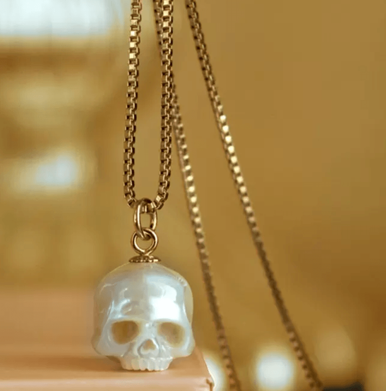 Pearlescent skull pendant on a gold box chain necklace, displayed on a warm-toned background.