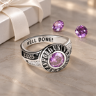 Close-up of personalized sterling silver graduation class ring featuring a purple birthstone, custom school name, 2025 engraving, and inside message. Displayed on an elegant beige marble surface with a white ribbon gift box and purple gemstones in the background.