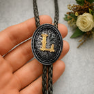Personalized bolo tie with gold letter L, black braided cord, and ornate silver-toned design, perfect custom gift.