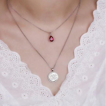 Double-layered necklace in sterling silver with a teardrop birthstone pendant and CZ monogram pendant, worn on a woman's neck with a white lace top.