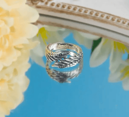 A silver ring with intricate feather designs engraved with the name "Andrea," reflected on a mirrored surface, surrounded by yellow flowers.