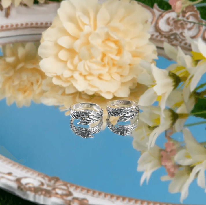 Two silver rings with intricate feather designs, engraved with the names "Andrea" and "Jason," reflected on a mirrored surface, surrounded by yellow flowers.