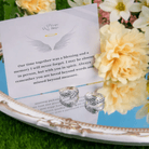 Two silver rings with feather designs, engraved with "Jason" and "Andrea," reflected on a mirror with a heartfelt message card, surrounded by yellow flowers.