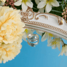 A silver ring with intricate feather designs engraved with the name "Andrea," reflected on a mirrored surface, surrounded by yellow and white flowers.