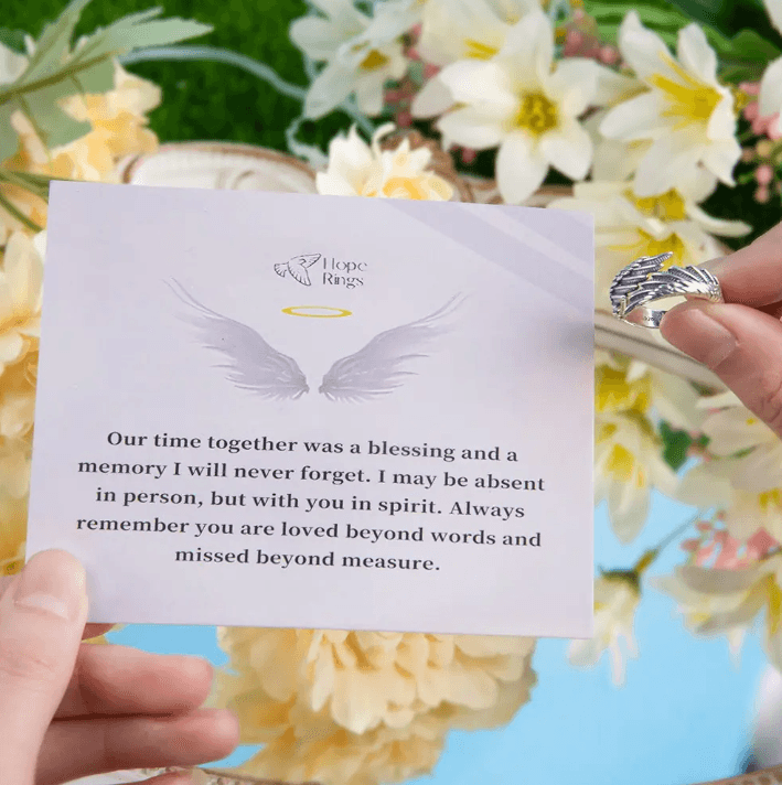 A hand holding a silver ring with feather designs next to a card with a heartfelt message, surrounded by yellow and white flowers in the background.