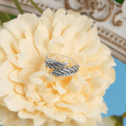 A silver ring with intricate feather designs engraved with the name "Andrea," placed on a large yellow flower with a mirror and decorative elements in the background.