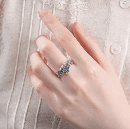 A hand wearing a silver ring with intricate feather designs, shown against a background of a light-colored, buttoned and lace-detailed blouse.