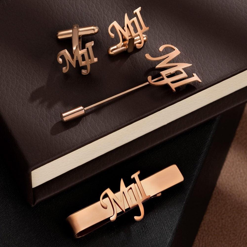 Rose gold monogrammed cufflinks, lapel pin, and tie bar set on a dark leather surface.