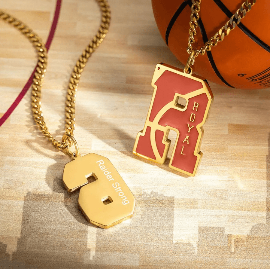 Two gold basketball-themed pendants on chains, one featuring the letter "R" with the name "Royal" and the other with the number "8" engraved with "Raider Strong," on a wooden floor next to a basketball.