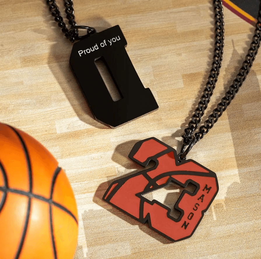 Two basketball-themed pendants on chains, one featuring the number "23" with the name "Mason" and the other with the letter "D" engraved with "Proud of you," on a wooden floor next to a basketball.