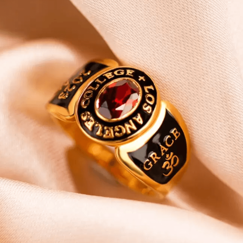 A gold college ring with a red gemstone, inscribed with "COLLEGE LOS ANGELES," the year "2024," and the name "GRACE" with an Om symbol, representing oneness and spirituality.