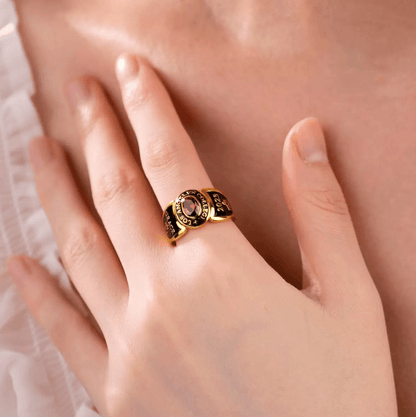 A gold college ring with a red gemstone, inscribed with "COLLEGE LOS ANGELES," "2024," and "GRACE" with an Om symbol, worn on a person's finger against their chest.