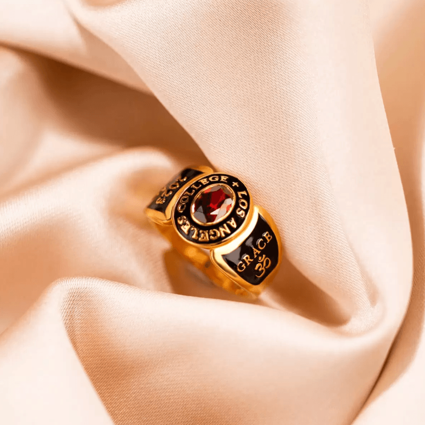 A gold college ring with a red gemstone, inscribed with "COLLEGE LOS ANGELES," "2024," and "GRACE" with an Om symbol, resting on beige fabric.