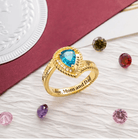 Gold personalized class ring with teal heart-shaped stone, engraved '2024, Kaplan Uni, Love, Mom and Dad' surrounded by gems.