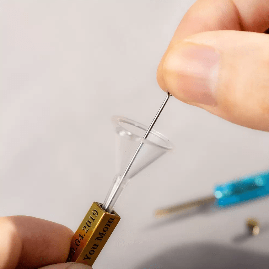 Filling a gold bar urn necklace with a funnel and miniature tool, engraved with a date and 'You Mom