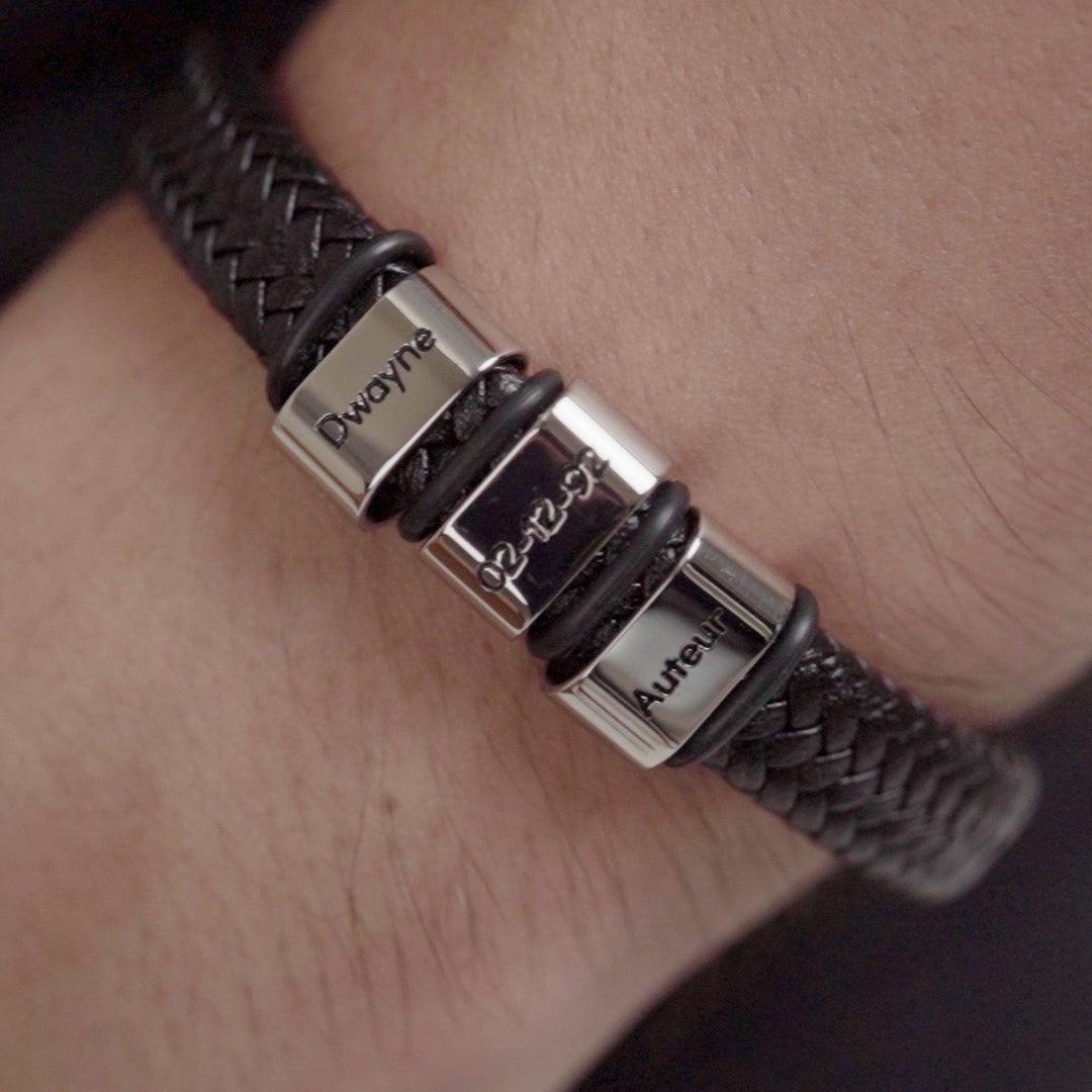 A close-up image of a wrist wearing a black braided leather bracelet with three metal beads. Each bead is engraved with the names "Dwayne," "Auteur," and a date "06-12-20."