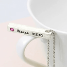 A silver necklace with a vertical bar pendant engraved with "Bianca 10.13.02" and a pink gemstone, draped over the rim of a white mug.