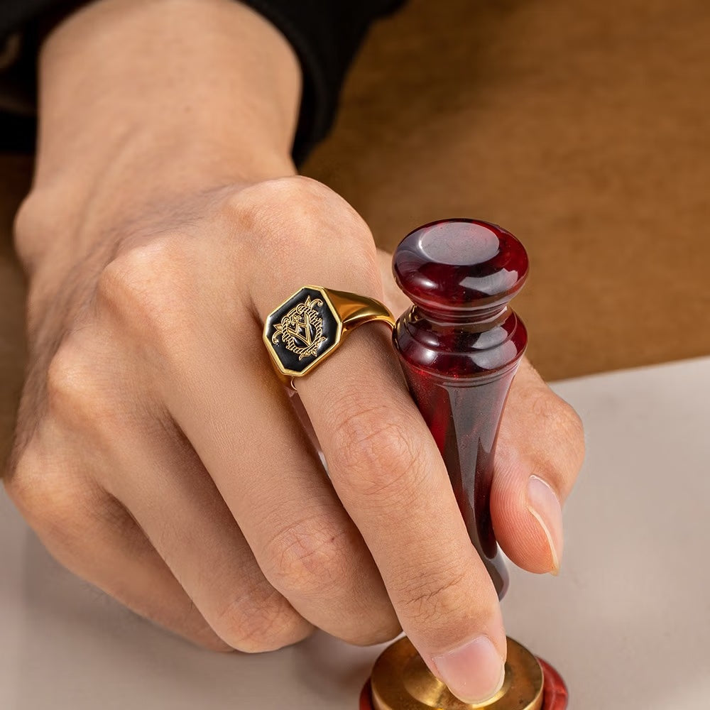 Gold tone custom double initials signet ring with black monogram face worn on a finger.