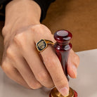Gold tone custom double initials signet ring with black monogram face worn on a finger.