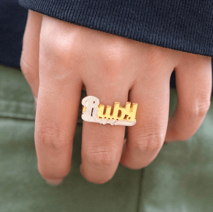 Gold and silver 'Ruby' name ring worn on the index finger of a hand against a green sleeve.