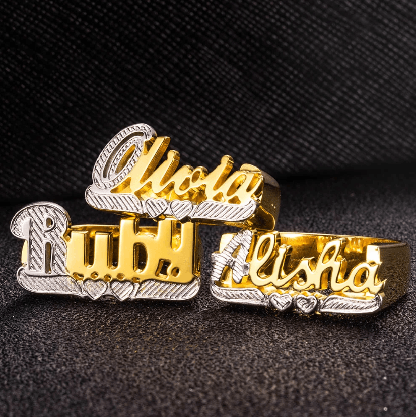 Gold and silver name rings displaying 'Olivia', 'Ruby', and 'Alisha', set against a textured black background.