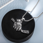 A silver necklace with a pendant shaped like a hockey glove holding a single hockey stick, resting on a black hockey puck.