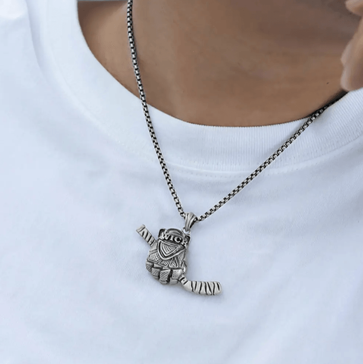 A silver necklace with a pendant shaped like a detailed hockey glove holding a single hockey stick, worn by a person in a white shirt.