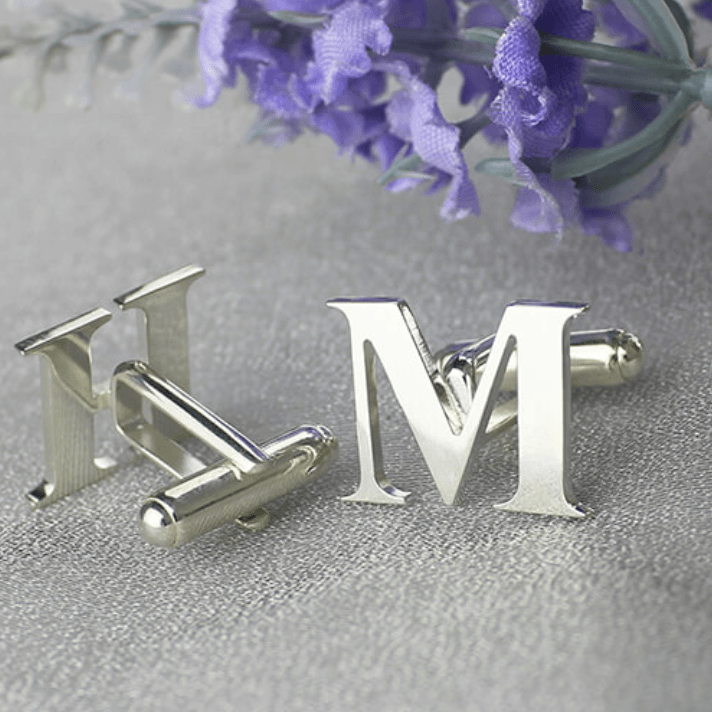 Silver initial cufflinks with the letters 'HM' on a textured grey surface with purple flowers in the background.