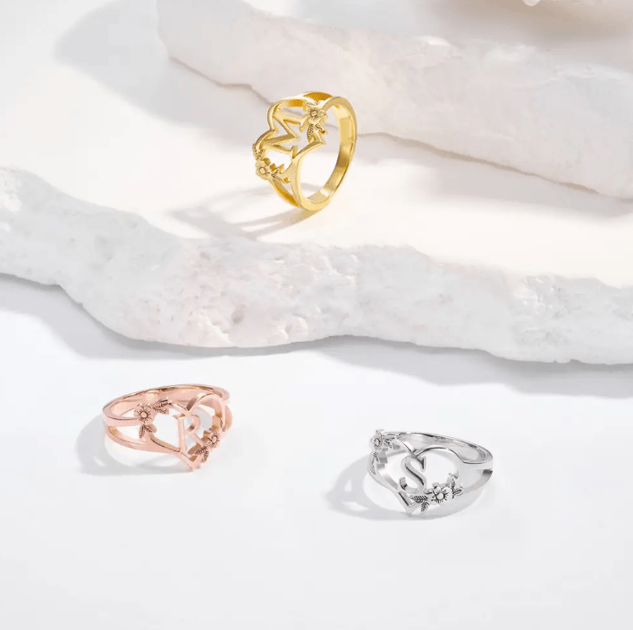 Three custom initial rings in gold, rose gold, and silver with heart and flower designs, displayed on a white surface.