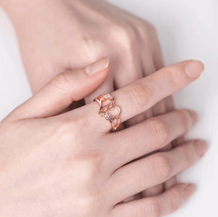 Rose gold ring with an initial letter encircled by a heart and flowers, worn on a woman's finger against a white background.