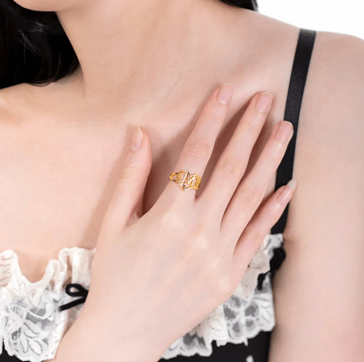 Gold initial ring with heart and floral accents on a woman's hand over a lace-trimmed top.