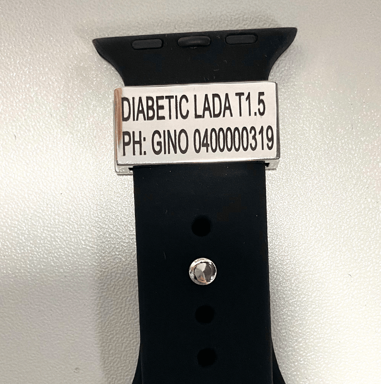 A silver medical tag on a black watch band with the inscription "DIABETIC LADA T1.5 PH: GINO 0400000319" displayed on a white background.