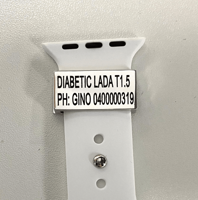 A silver medical tag on a white watch band with the inscription "DIABETIC LADA T1.5 PH: GINO 0400000319" displayed on a white background.