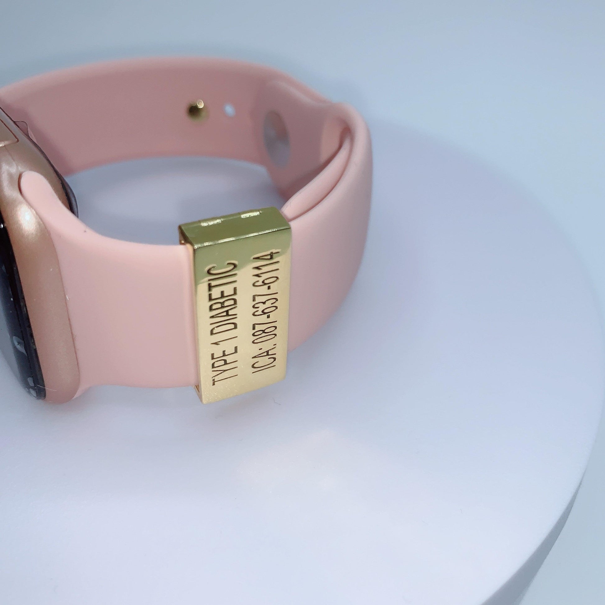 A gold medical tag on a pink smartwatch band with the inscription "TYPE 1 DIABETIC ICA: 087-637-6114" displayed on a white background.