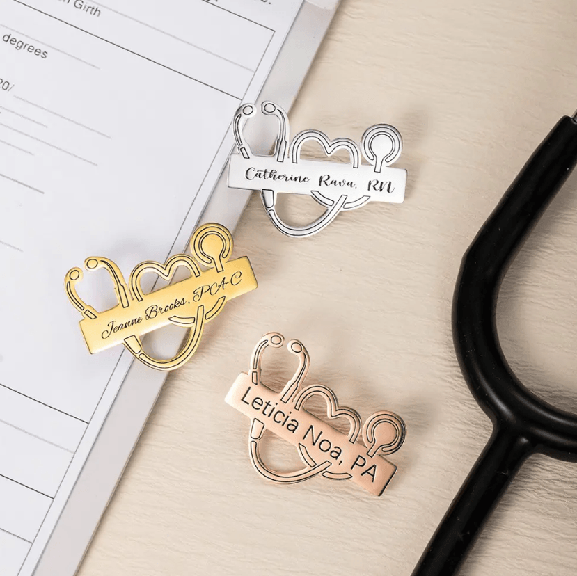 Three Personalized Medical School Graduation Lapel Pins in gold, silver, and rose gold with custom stethoscope designs, displayed next to a stethoscope and paperwork.