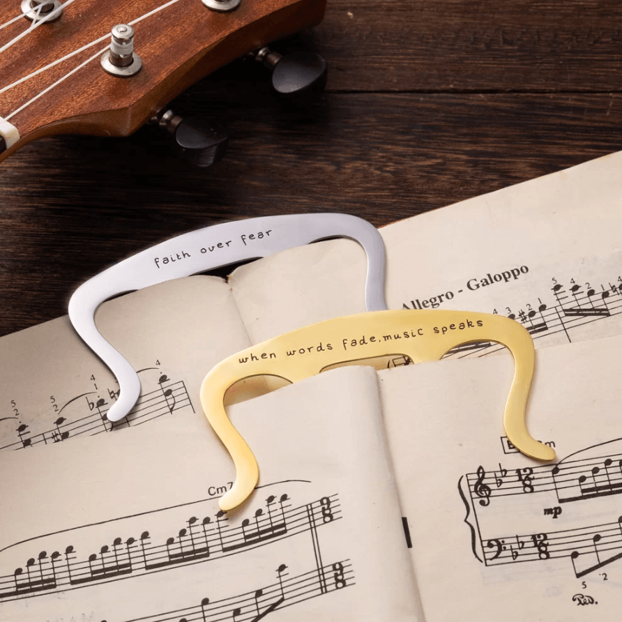 Two music book holder clips with inscriptions on sheet music next to a ukulele.