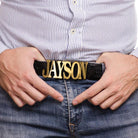 Man in striped shirt pointing to a black belt with custom gold buckle spelling 'JAYSON' against a denim jeans background.